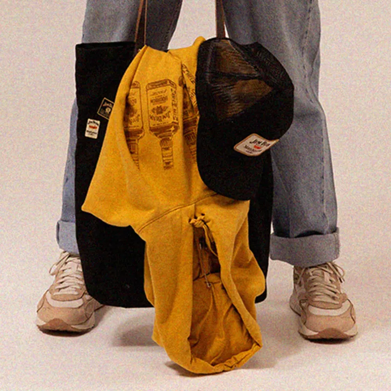 Person standing with a yellow hoodie and a black tote bag, wearing denim jeans and beige sneakers.