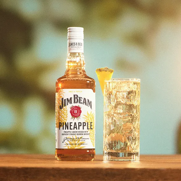 A bottle of Jim Beam Pineapple whiskey next to a tall glass filled with a refreshing cocktail and garnished with a pineapple slice.