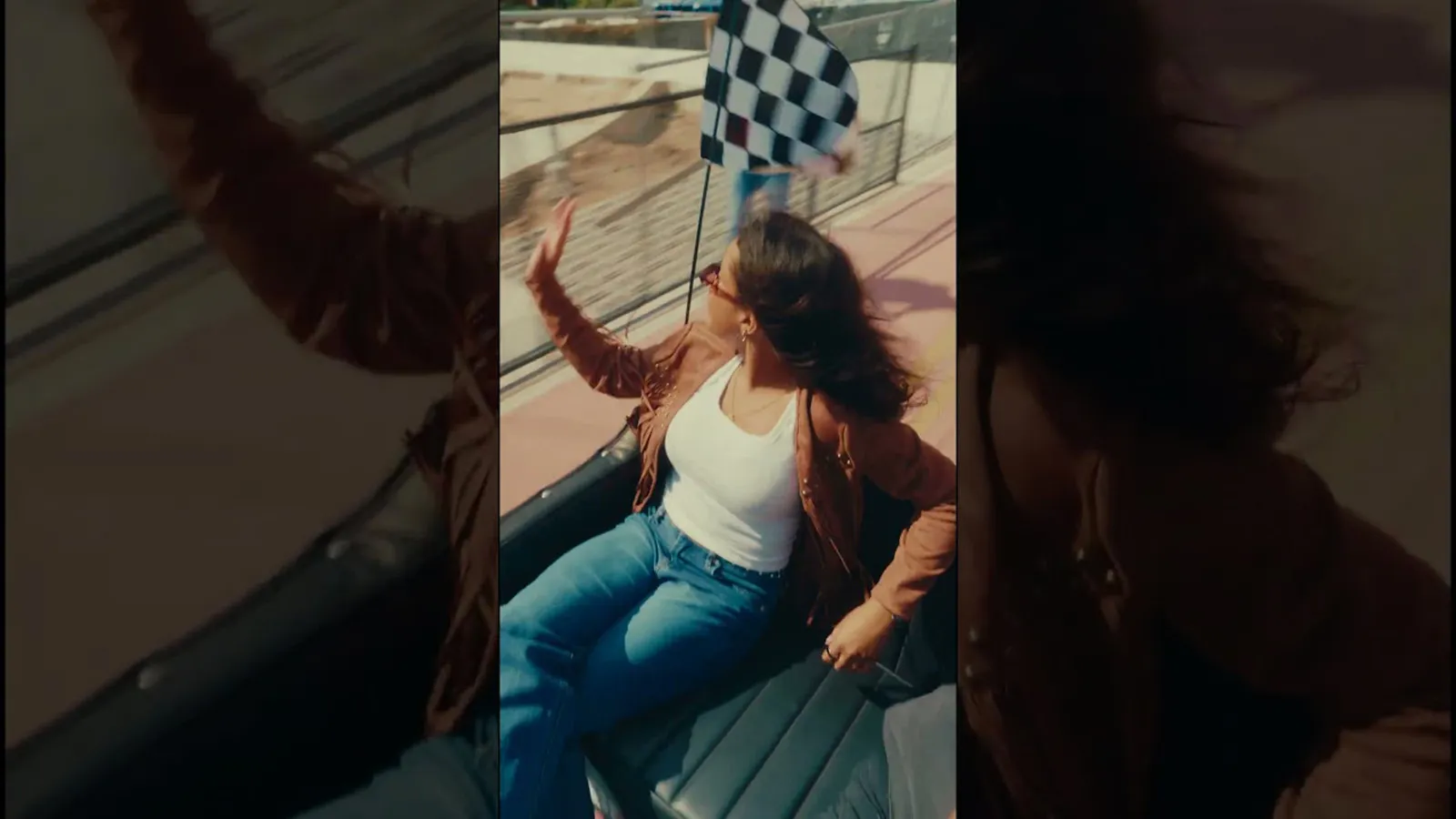 A woman in sunglasses joyfully waves from a car while a person nearby holds a checkered flag, signaling excitement and celebration.