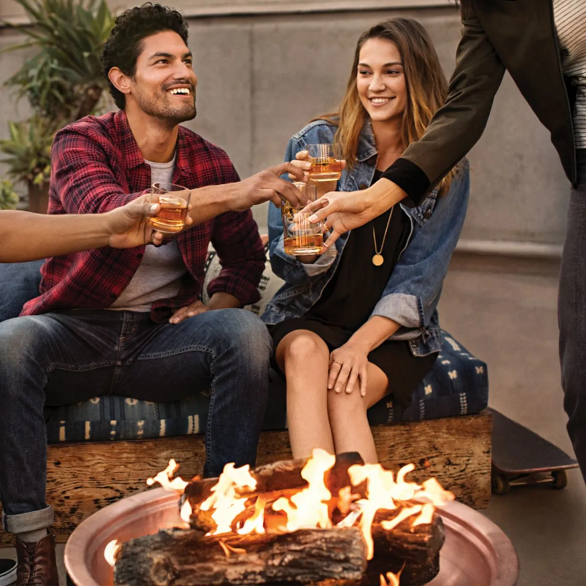 A group of friends toasting with glasses around a fire pit, enjoying a casual gathering in a cozy outdoor setting.