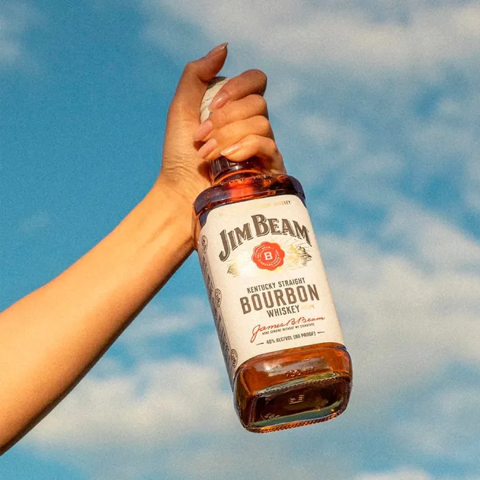 Hand holding a bottle of Jim Beam Kentucky Straight Bourbon Whiskey up against a bright blue sky.