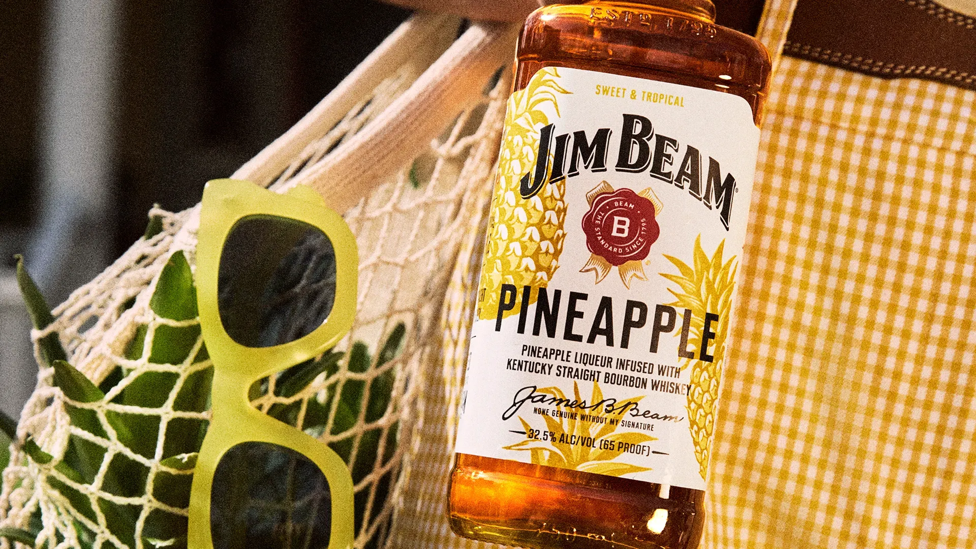Jim Beam Pineapple bottle styled with sunglasses and a net bag—tropical vibes for mixing a Bourbon Colada.