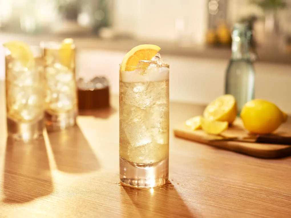 Whiskey & Soda Highball over ice with an lemon slice garnish on a bar, with citrus and soda in the background.