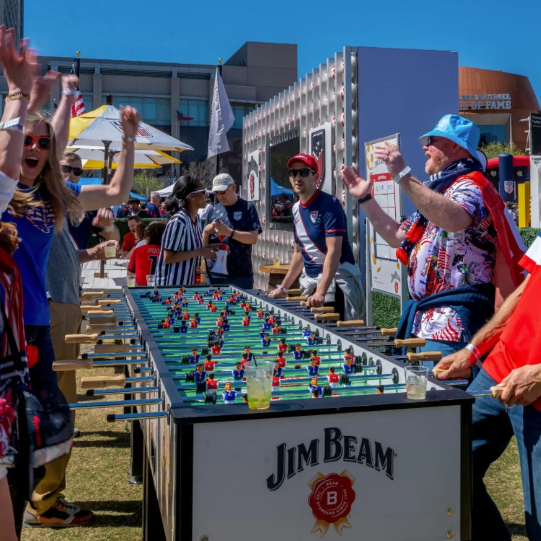 Fans cheer around a foosball table at an outdoor soccer festival under a bright blue sky.